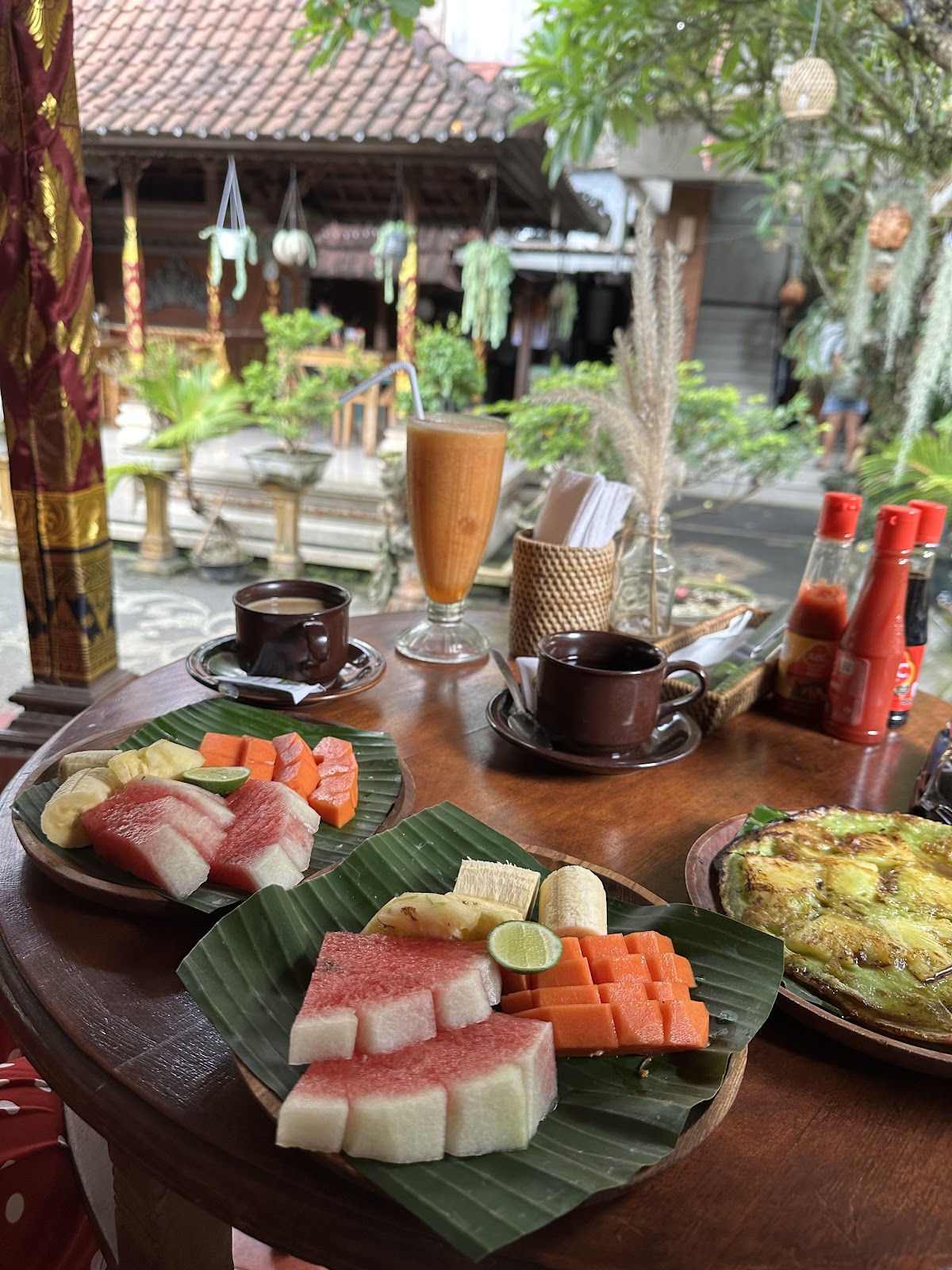 Elegant dining setup at a traditional Balinese homestay with fresh sashimi platters on banana leaves, traditional coffee service, and a serene courtyard view with tropical architecture and lush vegetation