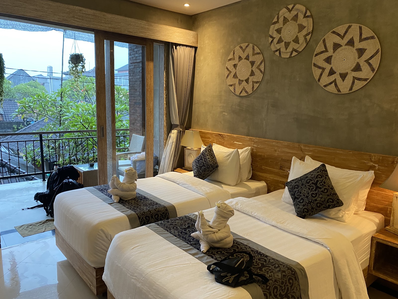 Twin bedroom with wooden headboards, white bedding, decorative patterned pillows, towel animals, and access to a balcony overlooking the city. Warm earth-toned walls decorated with woven circular wall art. Modern and comfortable accommodation setup.