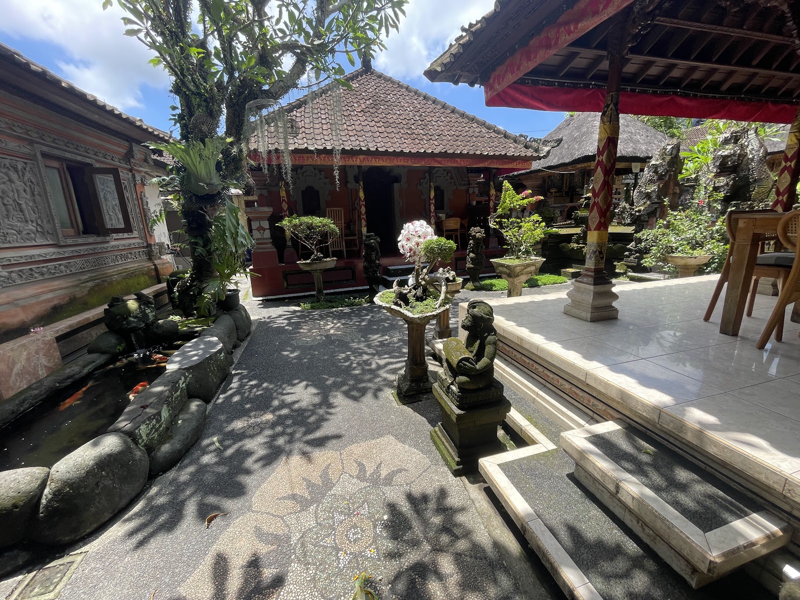 Traditional Balinese courtyard with red-roofed buildings, stone statues, lush tropical vegetation, and decorative umbrellas creating an authentic cultural atmosphere