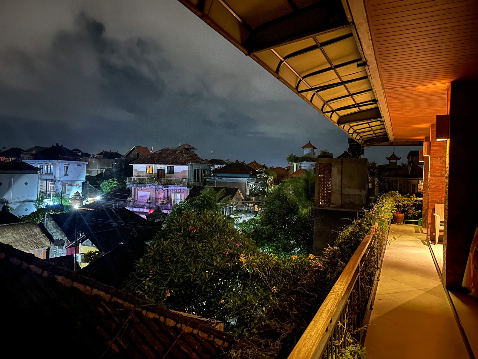 Evening twilight view from a balcony or terrace of a Southeast Asian town with traditional architecture, illuminated buildings, lush vegetation, and a covered walkway in foreground. Dramatic sky with moody lighting.